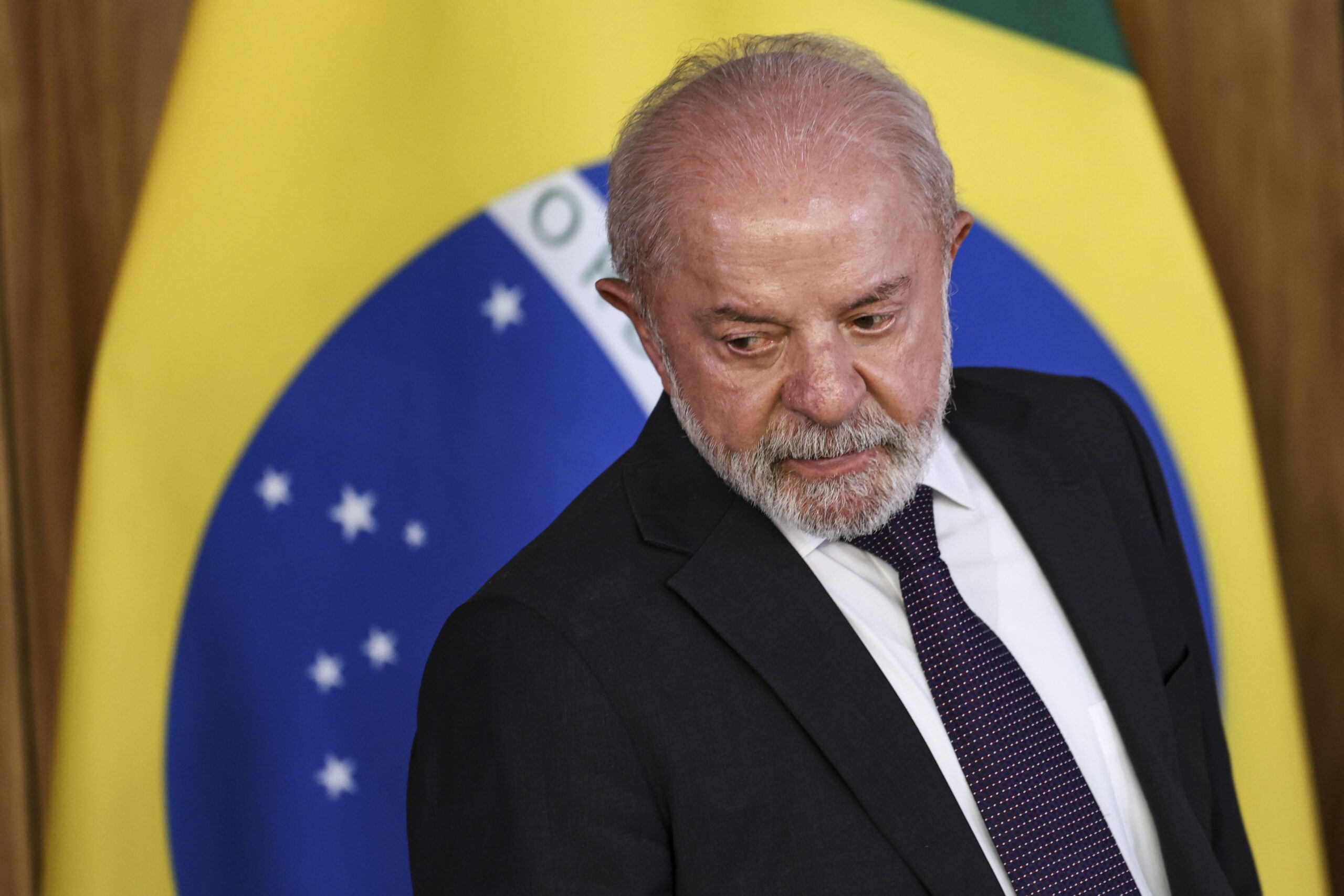 A imagem mostra o presidente Luiz Inácio Lula da Silva vestindo um terno preto, com camisa branca e gravata escura. Ele olha para o lado baixo-esquerdo da imagem. Ao fundo, é possível ver a bandeira do Brasil desfocada