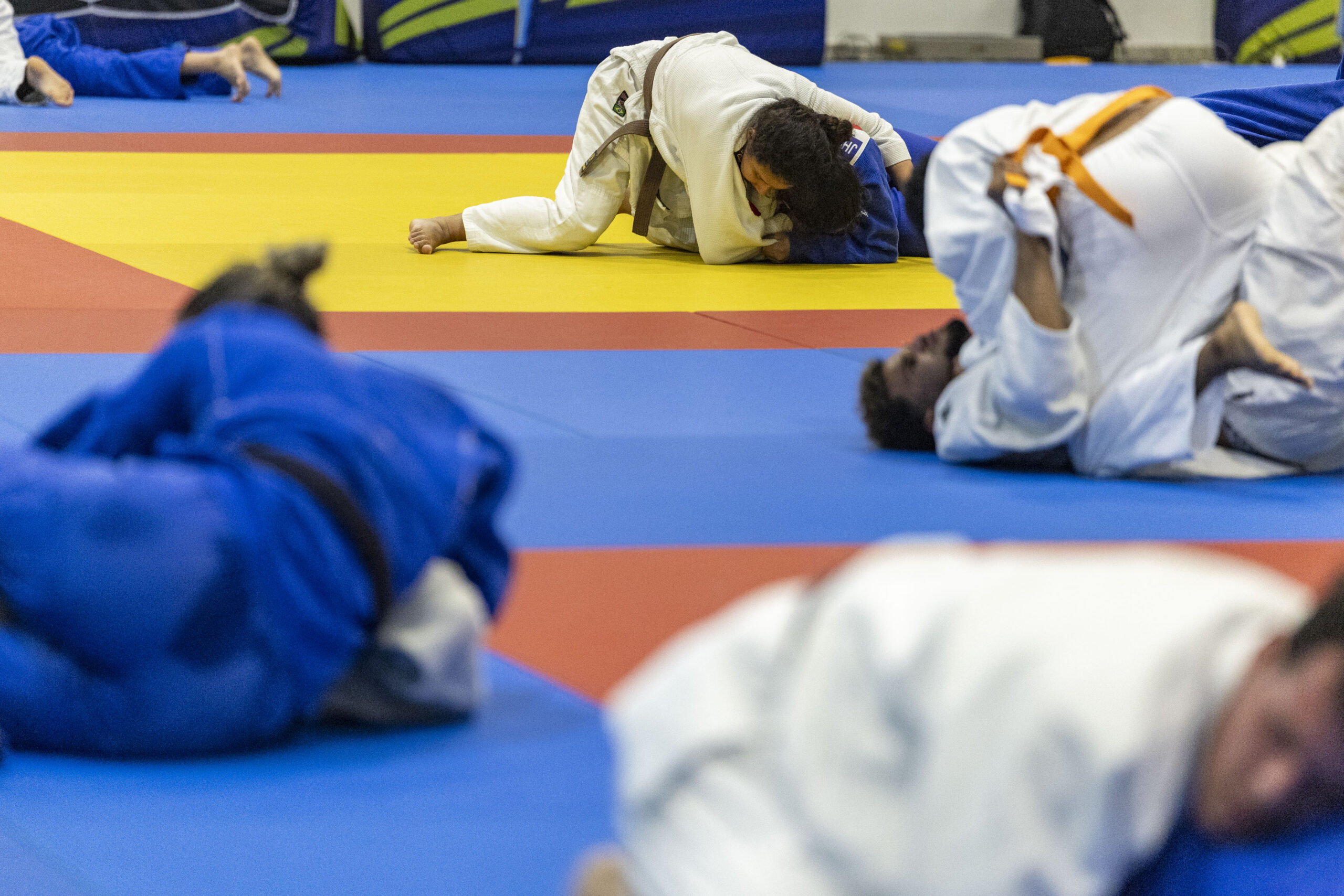 A imagem apresenta um treino de judô, com participantes usando quimonos azuis e brancos. Alguns estão com faixas pretas, enquanto outros usam faixas cinzas e amarelas. O tatame é das cores azul, vermelho e amarelo.