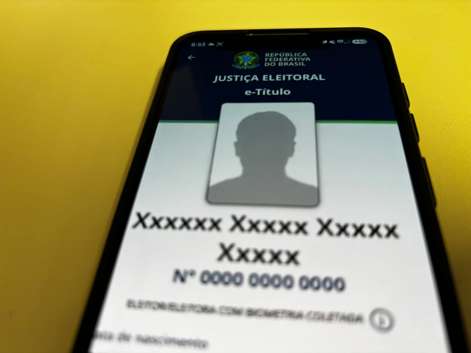 Fotografia aproximada de um telefone celular sob um fundo amarelo, exibindo o app e-Título da Justiça Eleitoral aberto, com uma silhueta cinza sem foto.