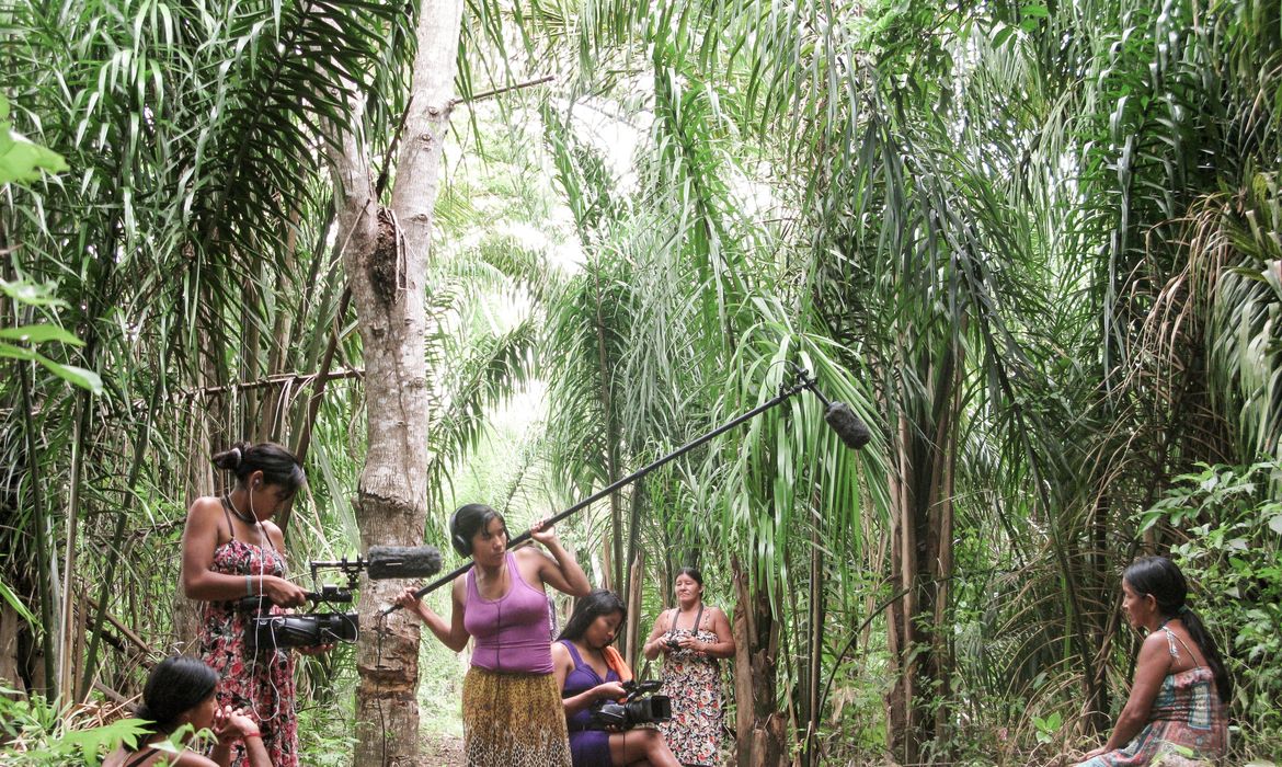 Mulheres indígenas realizando a gravação de um filme em meio à floresta. À esquerda, uma mulher usa fone de ouvido e segura uma câmera de vídeo, apontada para o centro da cena. Ao lado dela, outra mulher segura uma vara com microfone (boom) erguida acima do grupo, também usando fone de ouvido. No centro, uma mulher opera outra câmera, filmando de perto. Outras duas mulheres observam a cena sentadas, uma à esquerda em primeiro plano e outra à direita. Ao fundo, vegetação densa de palmeiras e árvores da floresta. 
