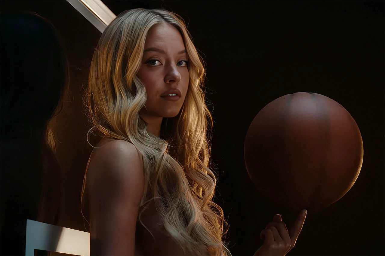 A atriz Sydney Sweeney, de cabelos loiros, segura uma bola de basquete com a ponta do dedo em uma cena escura da campanha publicitária da Novig