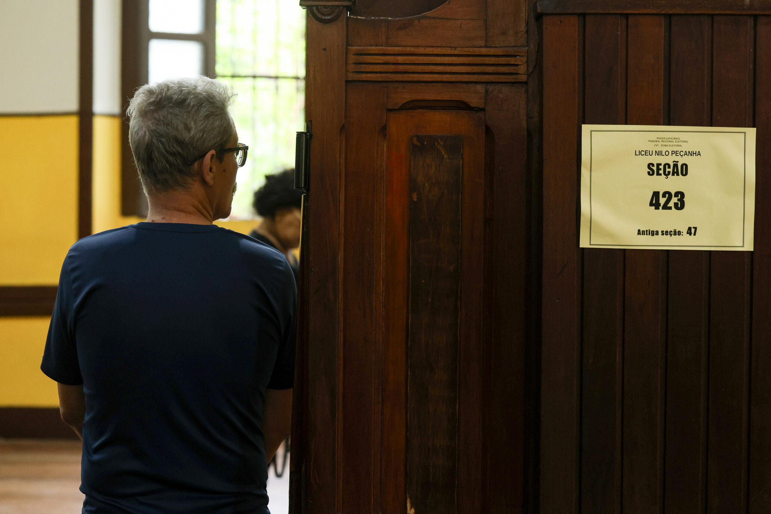 Pessoa idosa esta encostada na porta de uma sala, ele está usando camisa azul escura, tem cabelos brancos e usa óculos e esta com a cabeça levemente virada para o lado direito. Na porta há um papel colado escrito "Liceu Nilo Peçanha, Seção 423, antiga seção 47".