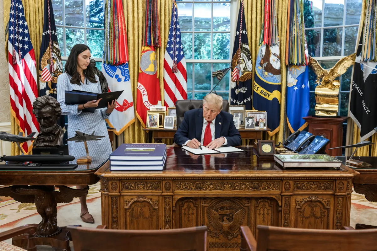 Donald Trump sentado à mesa de madeira na Sala Oval, a assinar um documento. O cenário de fundo inclui grandes janelas e várias bandeiras enfileiradas, como a dos Estados Unidos, do Corpo de Fuzileiros Navais, da Marinha e da Força Espacial.