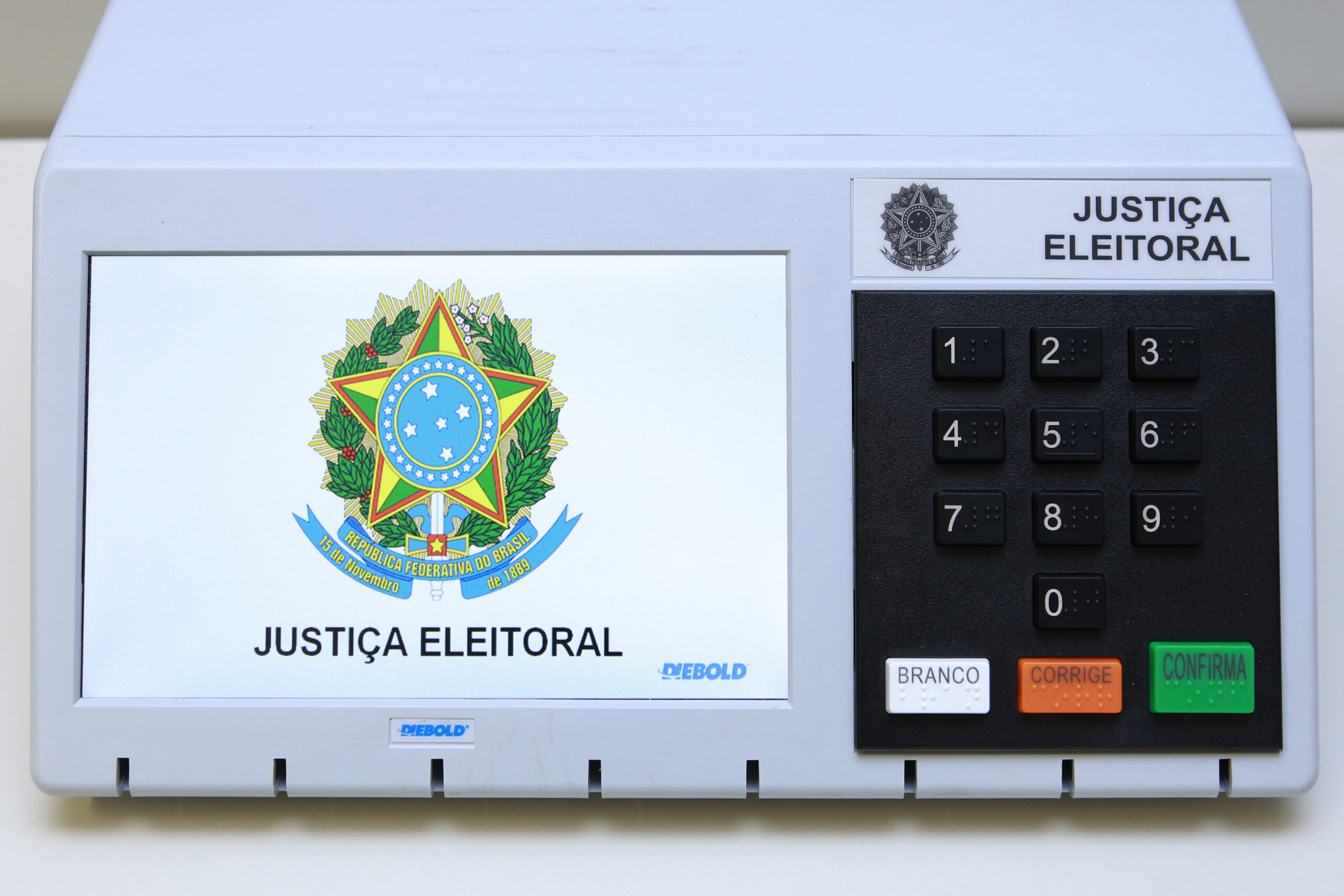 Urna eletrônica brasileira com tela exibindo o brasão da República e o teclado numérico com as teclas Branco, Corrige e Confirma.