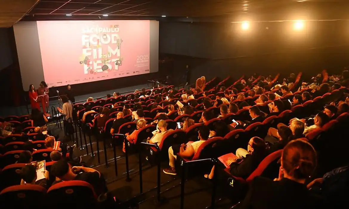 Plateia lotada em uma sala de cinema com poltronas vermelhas, assistindo à projeção da tela com o logotipo "São Paulo Food Film Fest 2025" em fundo rosa. No canto inferior esquerdo, duas pessoas em pé perto do palco, uma delas vestida de vermelho segurando um microfone, parecem apresentar o evento. A sala está com luzes baixas e o público, de costas para a câmera, observa a tela.